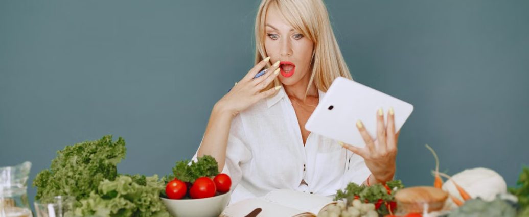 A surprised woman looking at a tablet surrounded by fresh vegetables and fruits, promoting healthy eating.