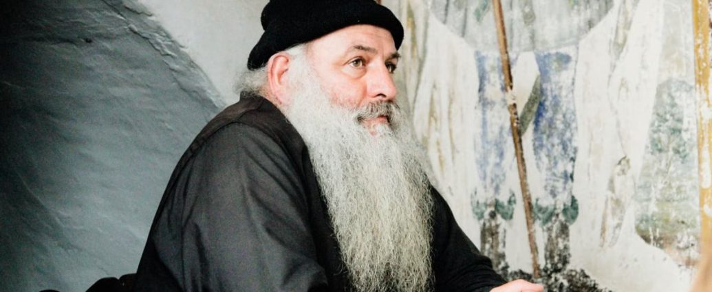 Elderly man with a beard wearing a beanie, seated inside a Greek monastery.