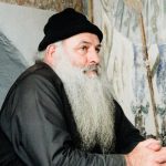 Elderly man with a beard wearing a beanie, seated inside a Greek monastery.