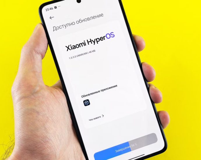 Hand holding smartphone showing Xiaomi HyperOS update screen on yellow background.